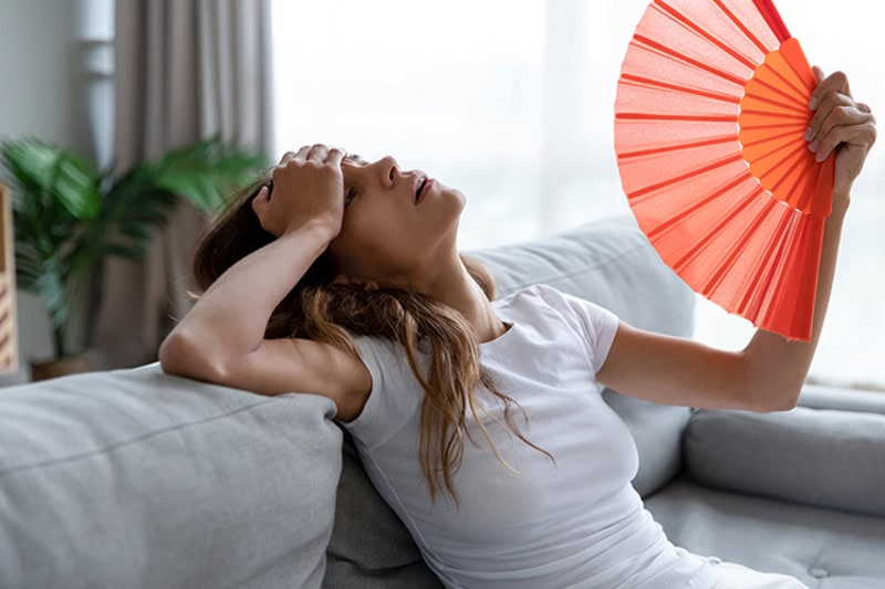 side view of an exhausted woman using a paper fan to cool down in her apartment.