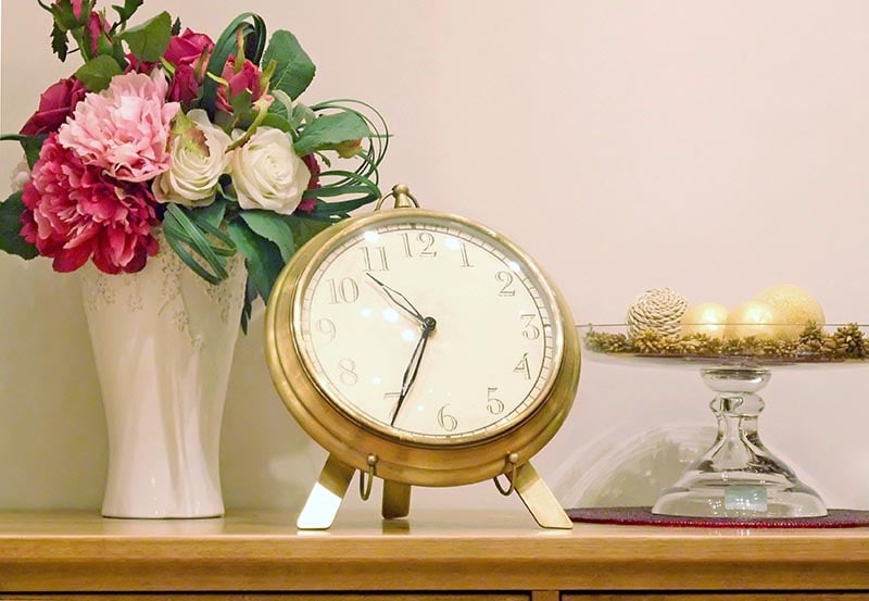 An ornate brass clock sits on a wooden tabletop between a white vase of pink and white flowers and a glass pedestal dish displaying decorative ornaments.