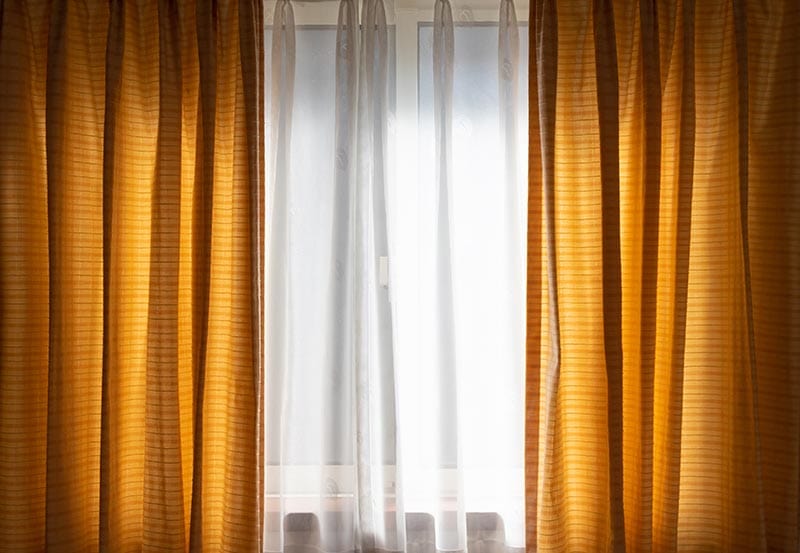 A window framed by warm, golden-brown or mustard-yellow drapes on either side. In the center, soft white sheer curtains filter bright daylight coming through the window panes.