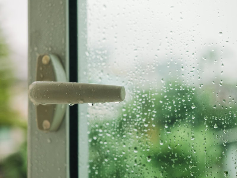 A white window latch handle covered in small water droplets. The windowpane is coated with rain or condensation, blurring a lush green outdoor background.
