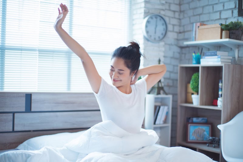 A young woman sitting in bed and stretching her arms with a smile upon waking up in the morning.